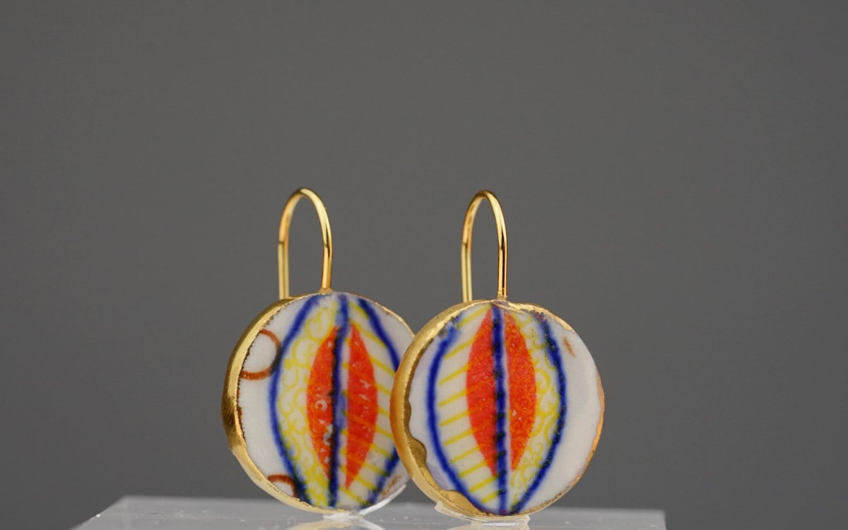 Die Vorderseite zeigt eine elegante elliptische Form in Rot, umrahmt von strahlendem Gelb und filigranen blauen Linien auf feinstem Porzellan. Die Rückseite ergänzt das Design mit einer verspielten Blattform in Orange und Blau, die dem Schmuckstück eine besondere Dynamik verleiht.